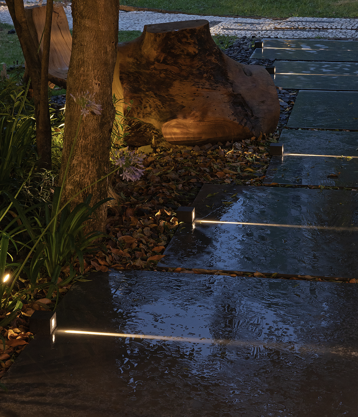 The image shows a pathway lit by Stella's Line bollards, with wet rectangular stones on the right. On the left, there are plant volumes, trees, flowers, and dry foliage.