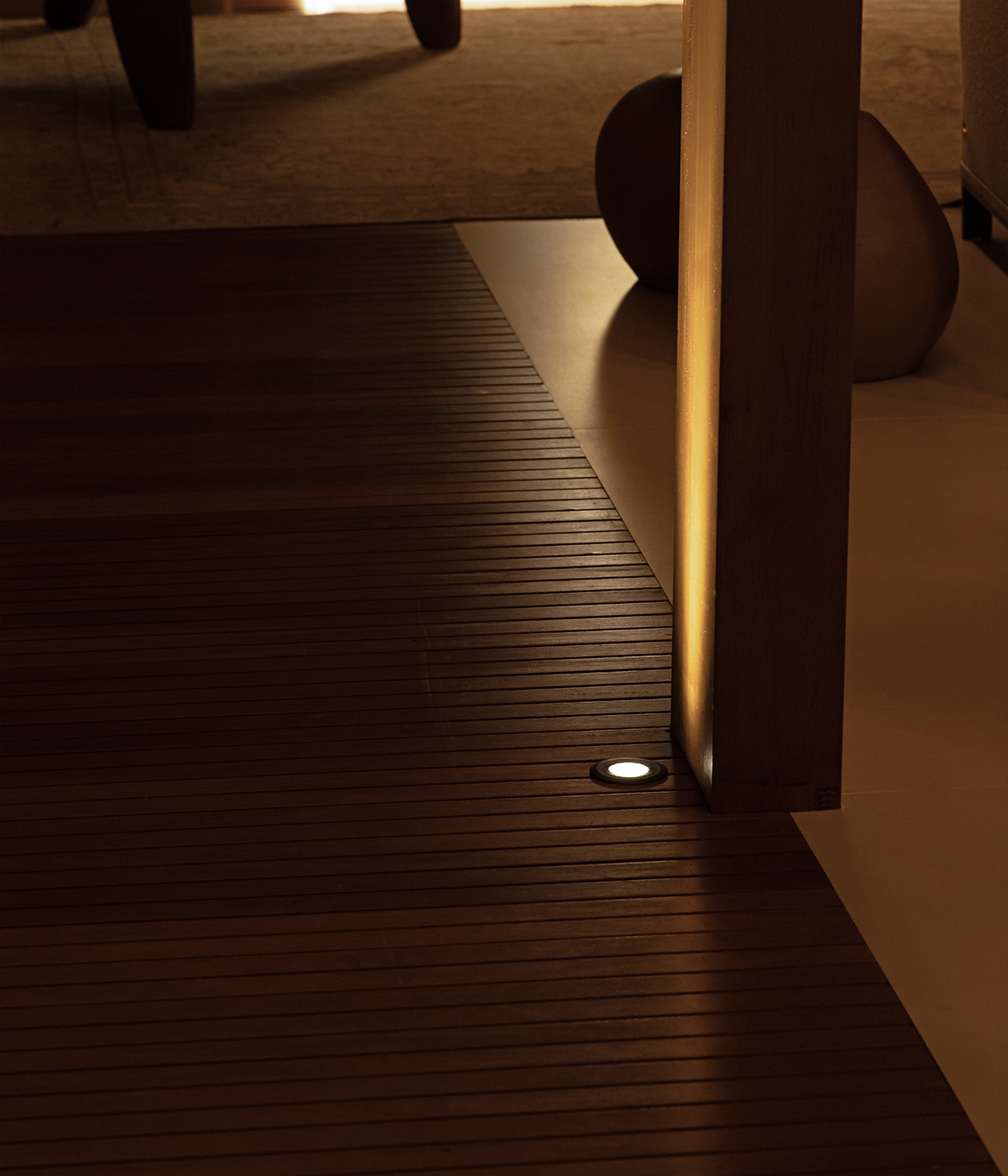 This image shows a darkened setting, with a transition from a deck on the left to a tiled floor on the right. Installed on the deck, an S.GROUND ground light illuminates a wooden pillar betwe