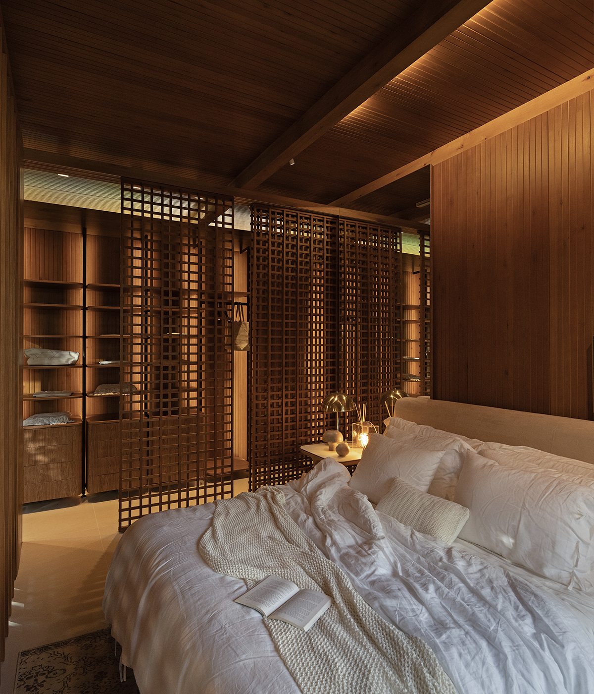 The image shows a bedroom and closet. In the foreground, a bed with white sheets and a book is lit from the ceiling by the Trace track system from Stella. In the background, the closet with w